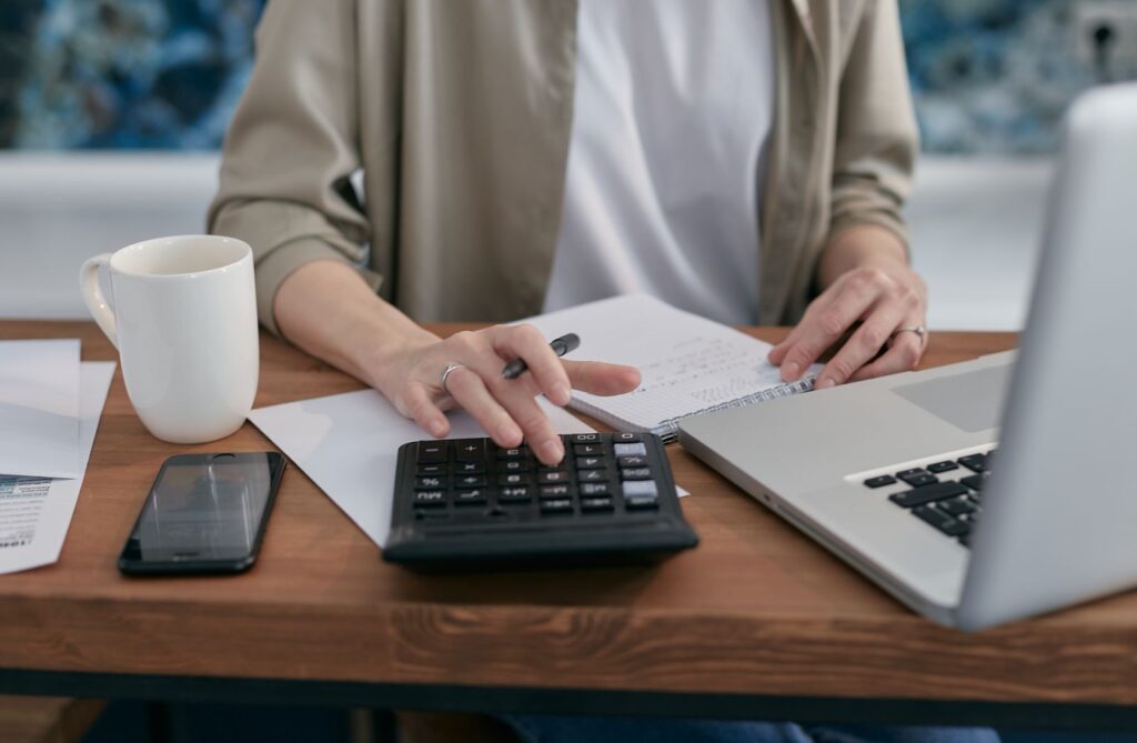 A person using a calculator and laptop while taking notes on business finances